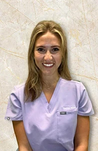 Dental hygienist Taylor in lavender scrubs smiling against marble wall backdrop.